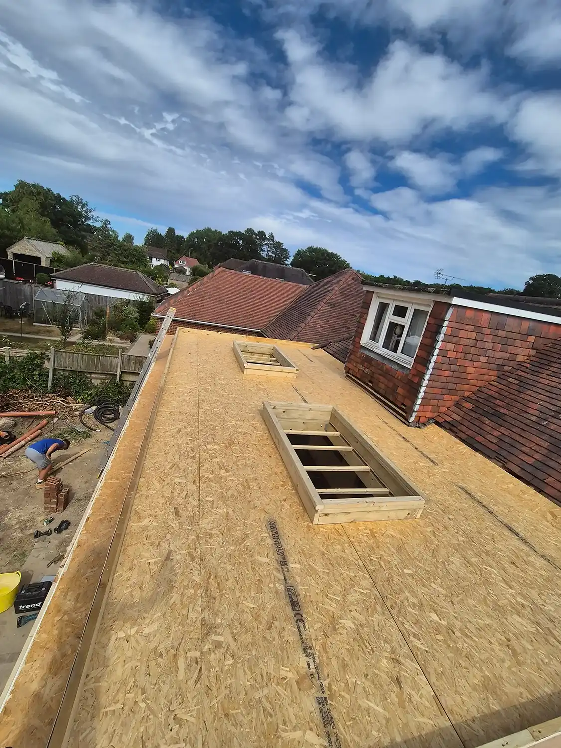 Flat wooden roof structure under construction with two framed window openings and a person working below.