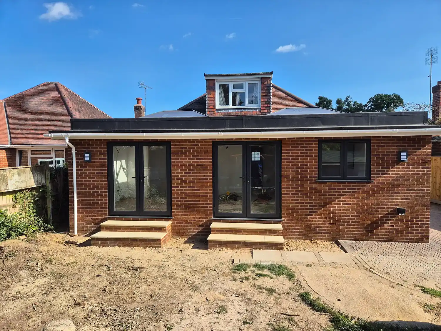Newly built brick house extension with two sets of black-framed glass doors and one window under blue sky.