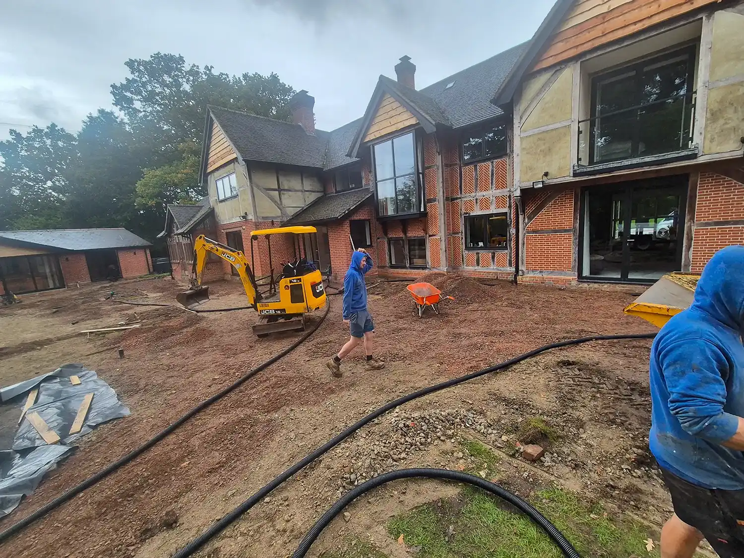 Construction site in front of a large brick and timber house with workers and a small excavator.