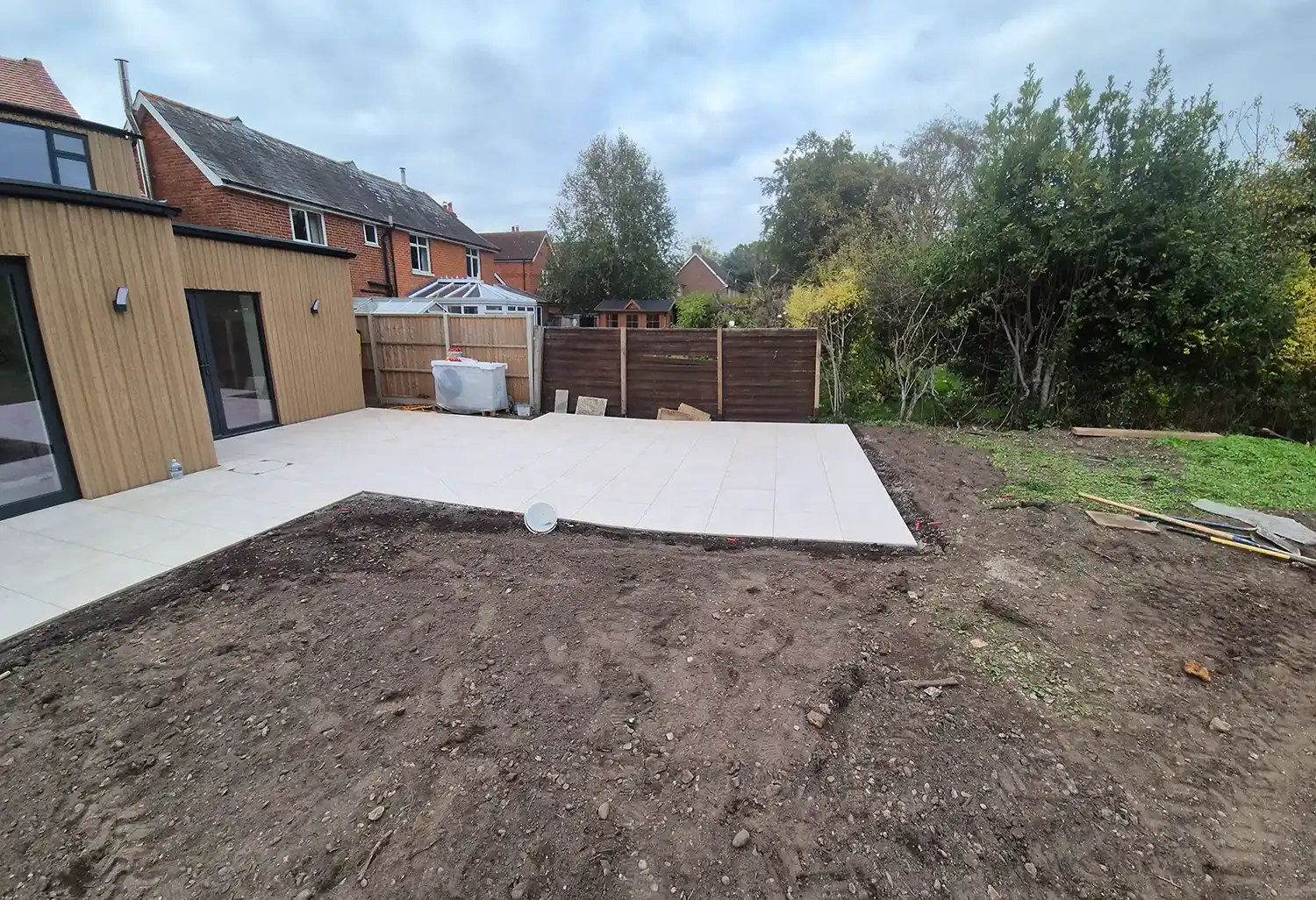 Backyard with newly installed tiled patio next to a house and bare soil area under cloudy sky.