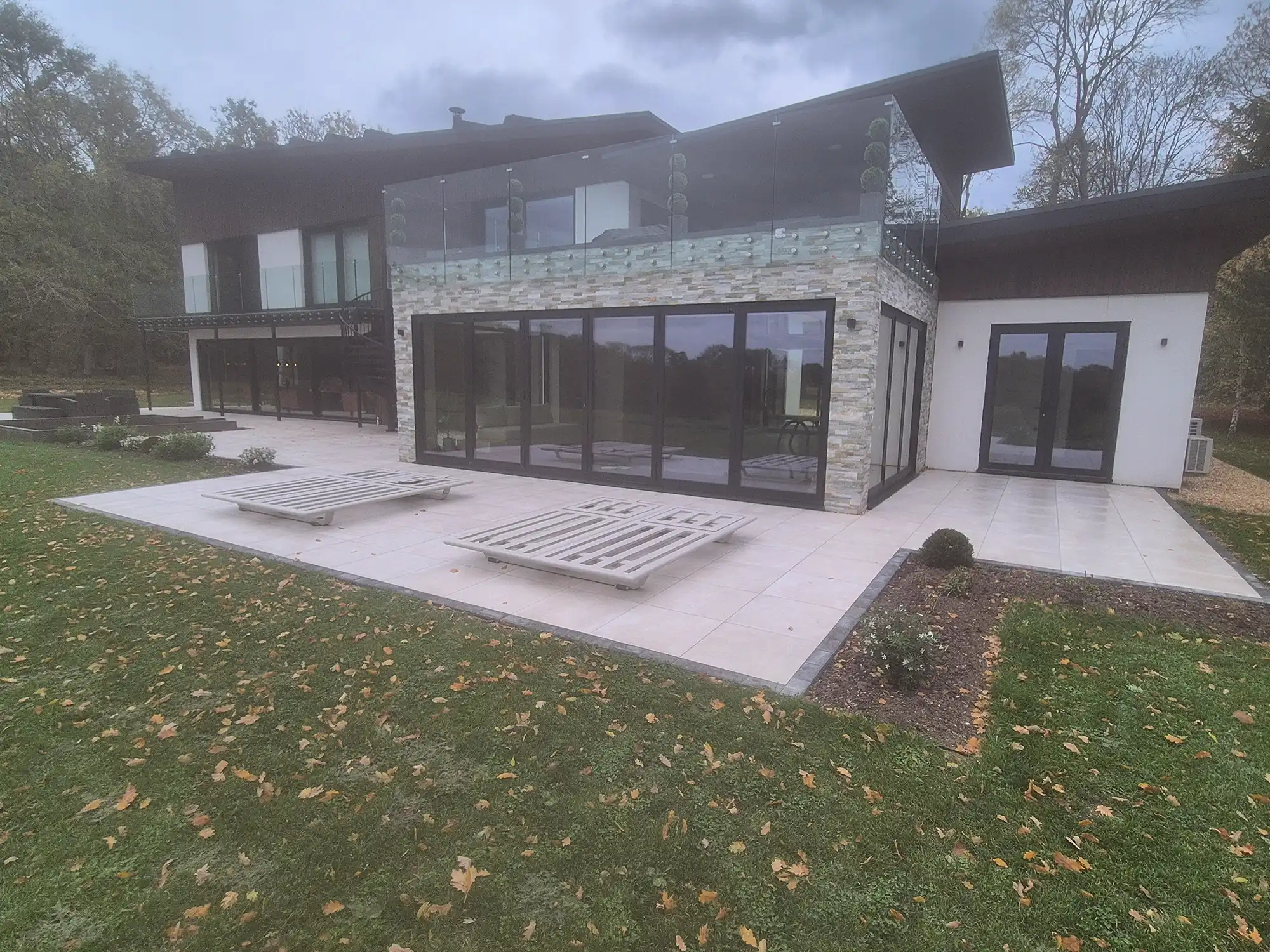 Modern house with large glass doors, stone facade, balcony with glass railing, and two lounge chairs on tiled patio.