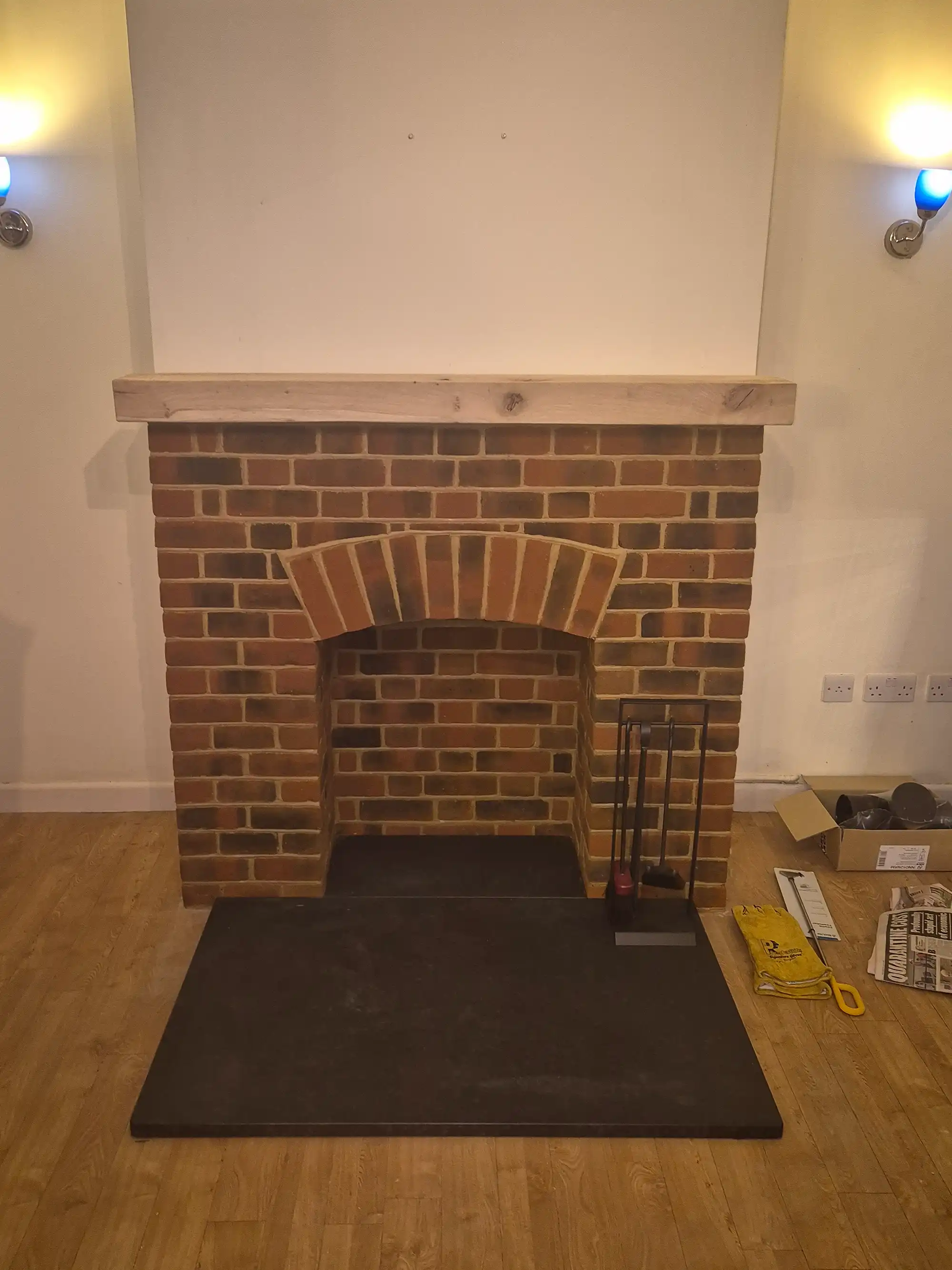 Empty brick fireplace with wooden mantle and black hearth on wooden floor under wall lights.