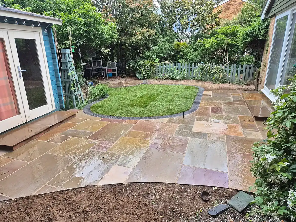 Back garden with wet stone patio, small grass lawn bordered by bricks, and dense green plants around.