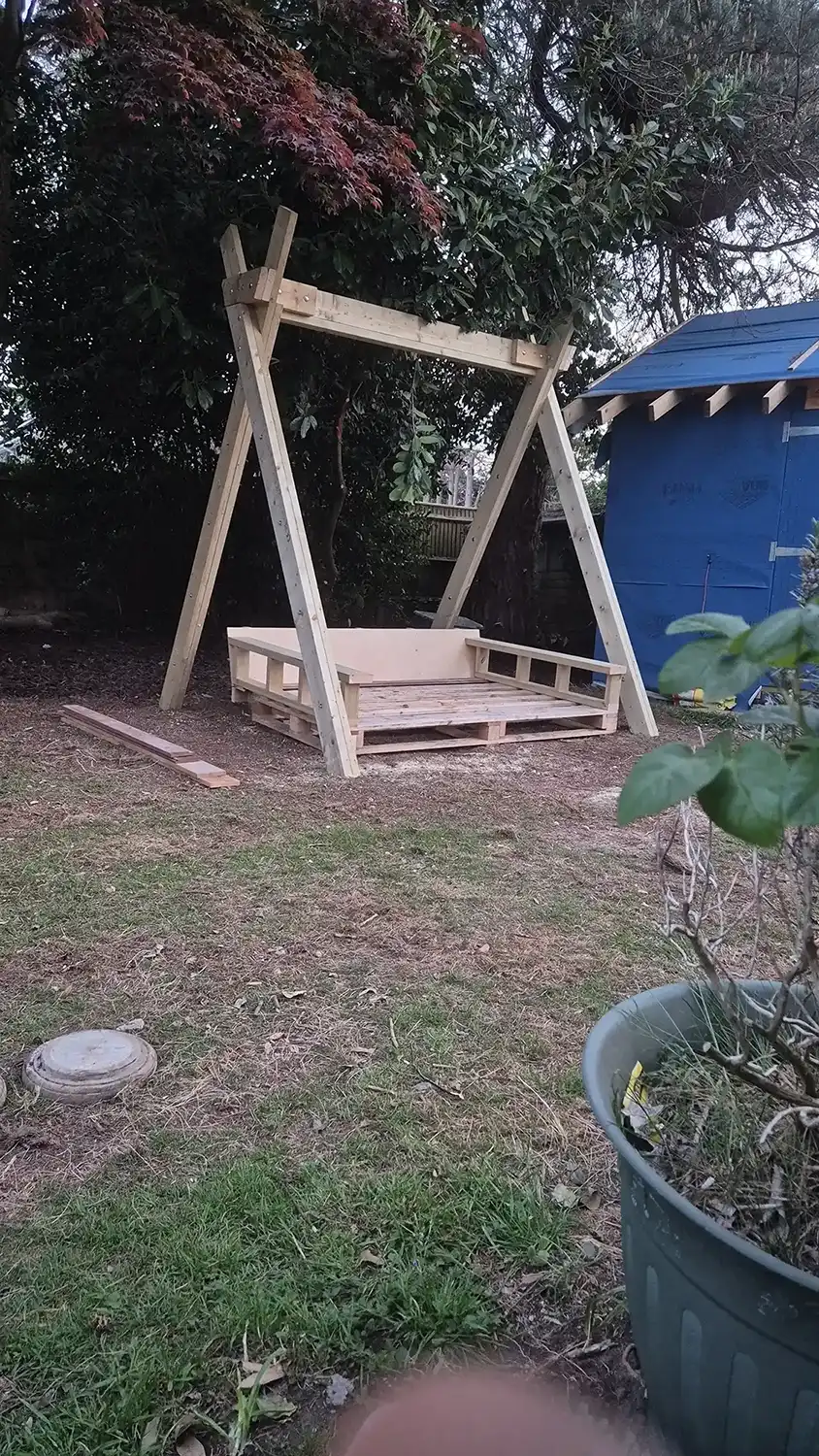 Unfinished wooden outdoor swing frame with a pallet seat set on grass in a back garden.