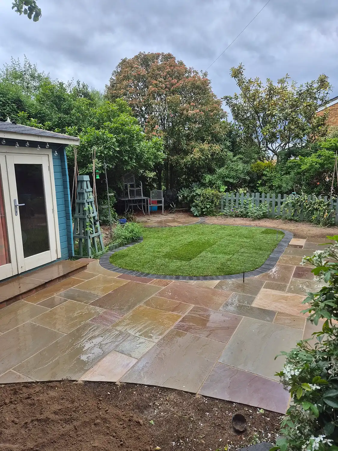 Back garden patio with wet stone tiles, small artificial grass area, surrounded by green trees and plants.