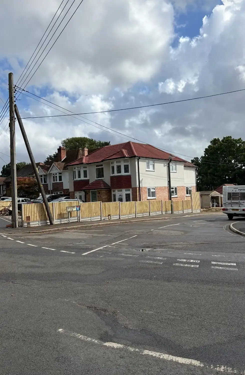 Two-story house with red roof, wooden fence, cars, utility poles, on a cloudy day at a street corner.