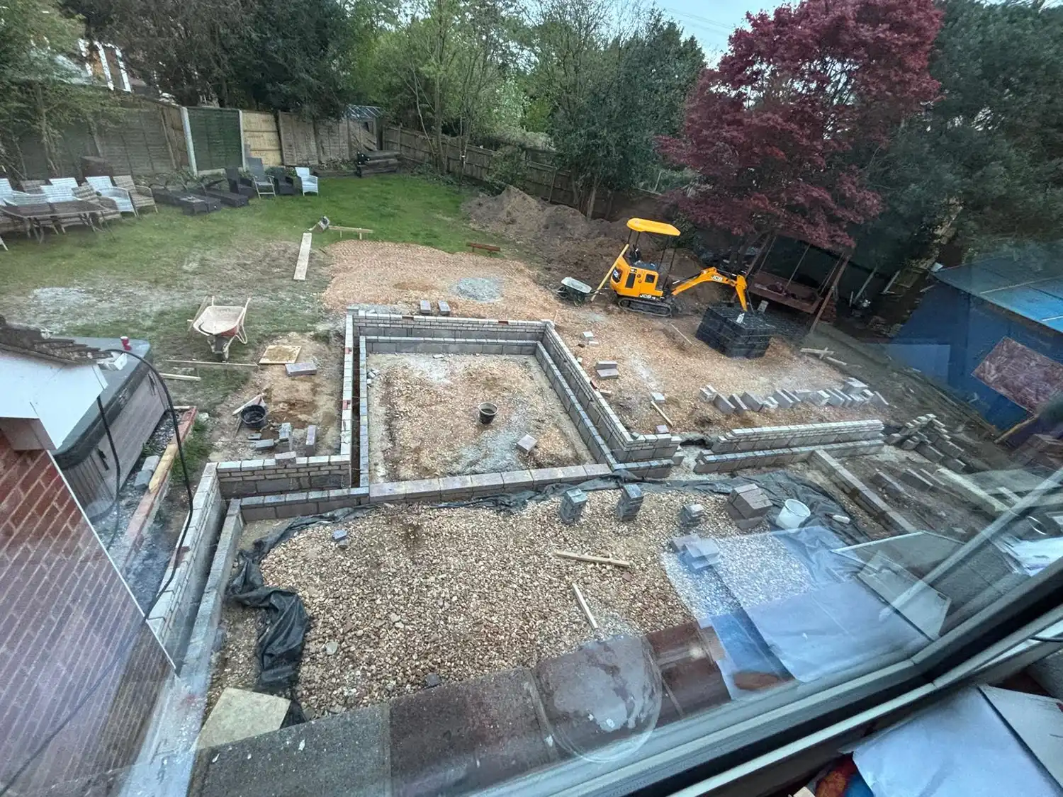Construction site in a backyard with foundation walls, gravel, a small excavator, and scattered bricks.