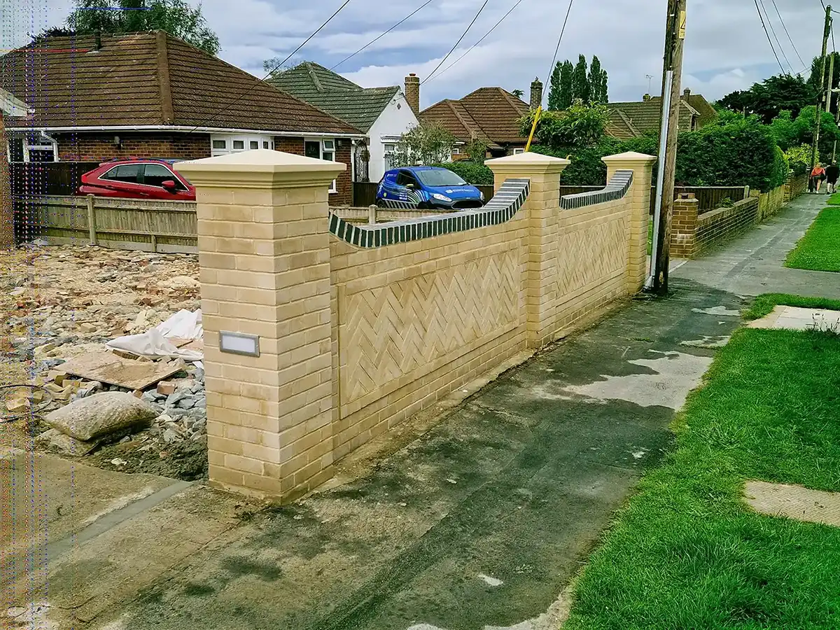 Light beige brick wall with decorative pattern running along a sidewalk in a suburban neighborhood.