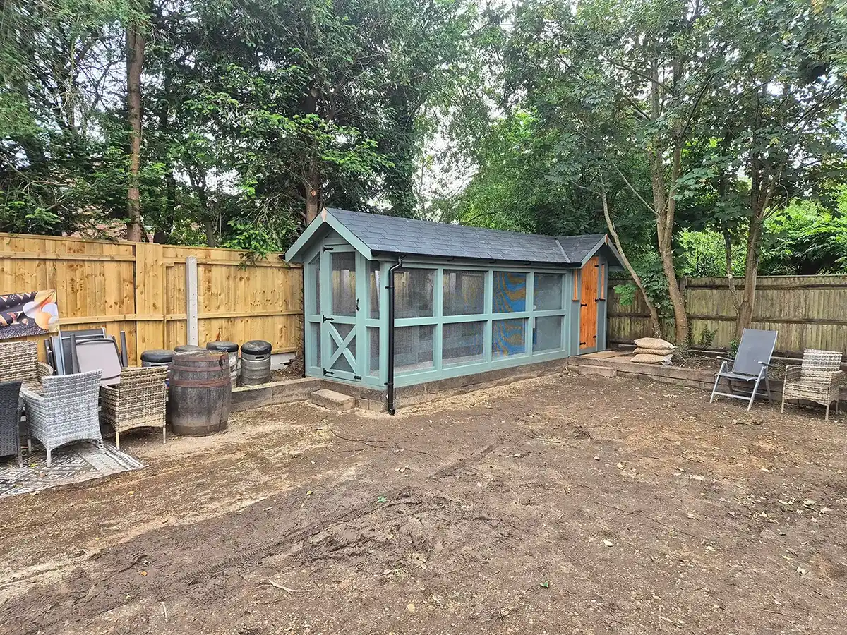 Blue and wood outdoor coop in backyard with wooden fence, chairs, and bare soil ground.