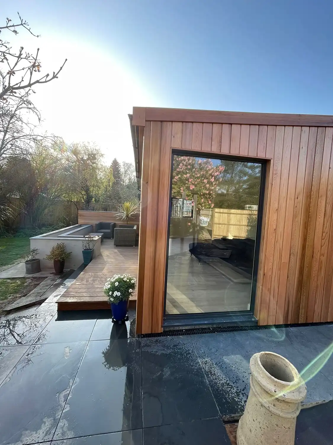 Modern wooden garden room with large glass door on wet stone patio and outdoor seating area.