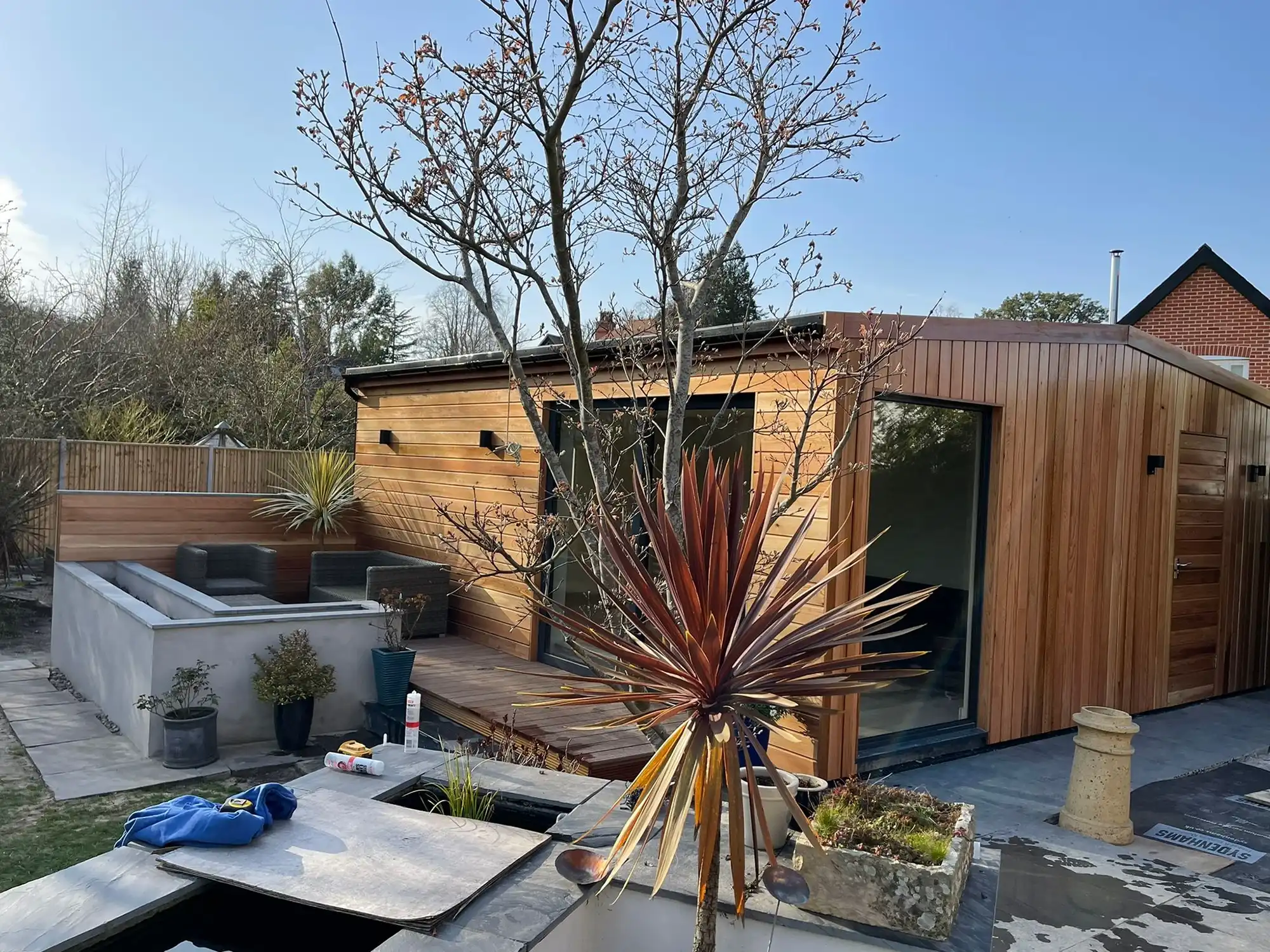 Modern wooden garden office with patio seating and plants under clear blue sky.