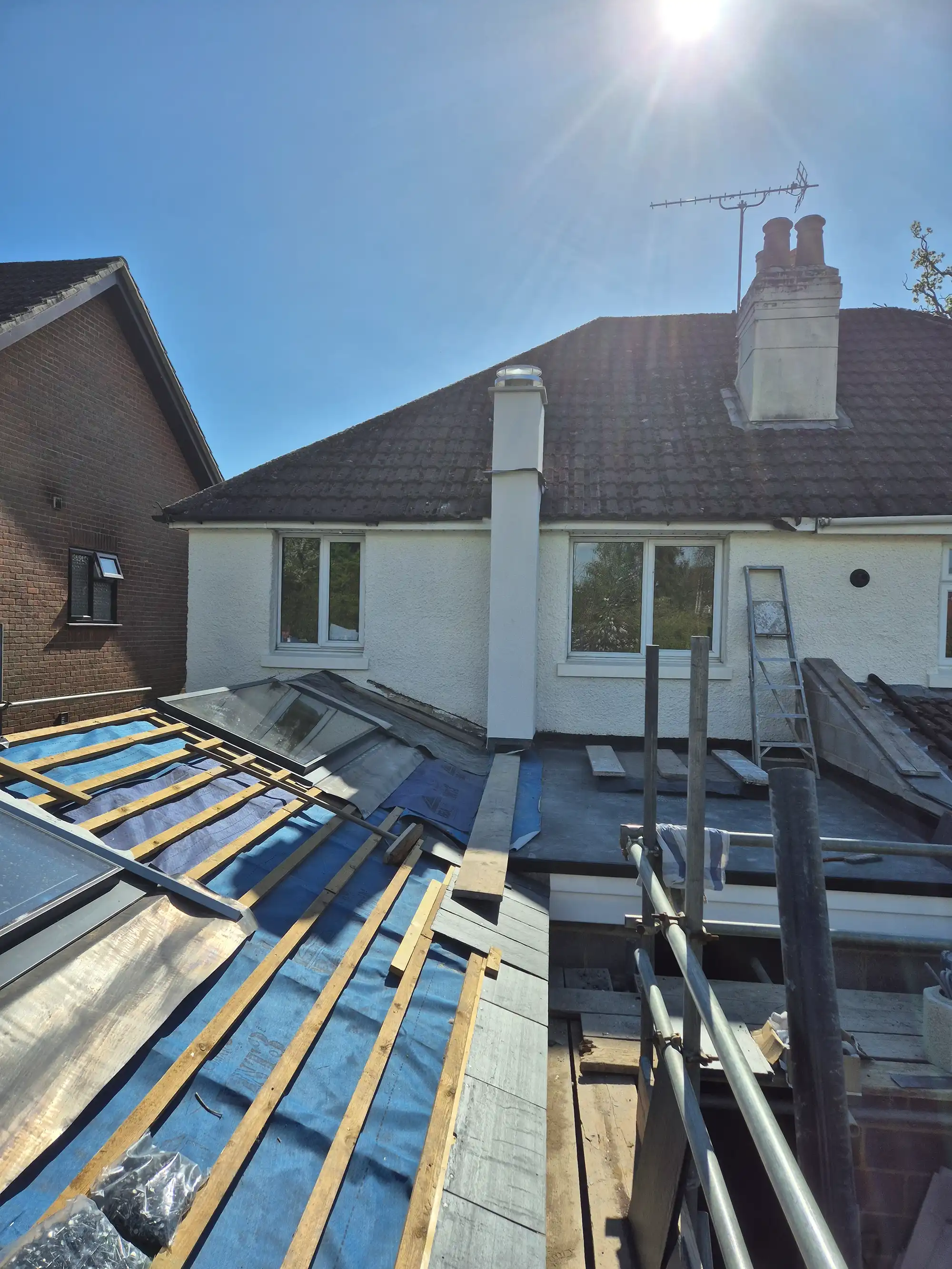 Roof under construction with blue underlay, wooden battens, scaffolding, and adjacent white house under bright sun.