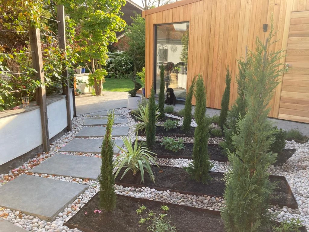 Modern garden with square stone path, tall slender shrubs, and a wooden building reflecting a black dog in the window.